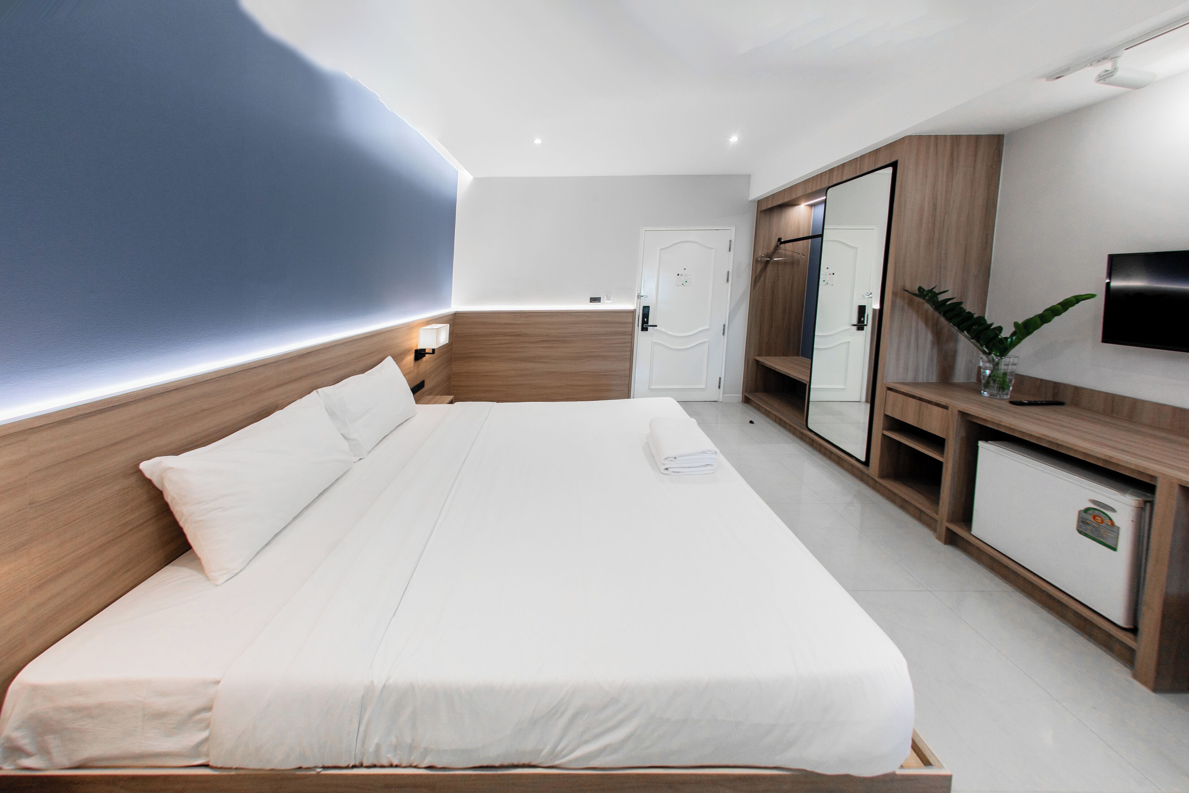Ein modernes Hotelzimmer mit einem großen Bett, einem Holz-Kopfteil, einem Kleiderschrank, einem Spiegel, einem Fernseher und einer kleinen Pflanze.