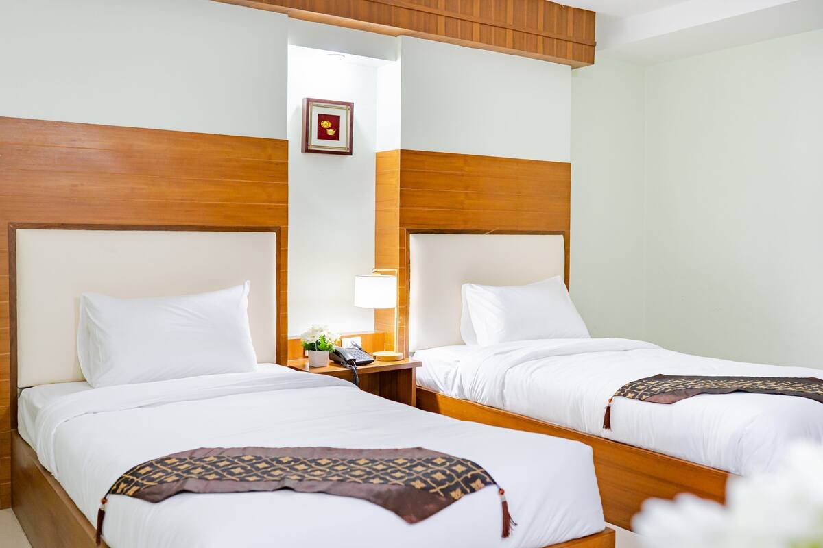 Een hotelkamer met twee bedden, houten hoofdborden, wit beddengoed, een nachtkastje met een lamp en een lijst aan de muur.