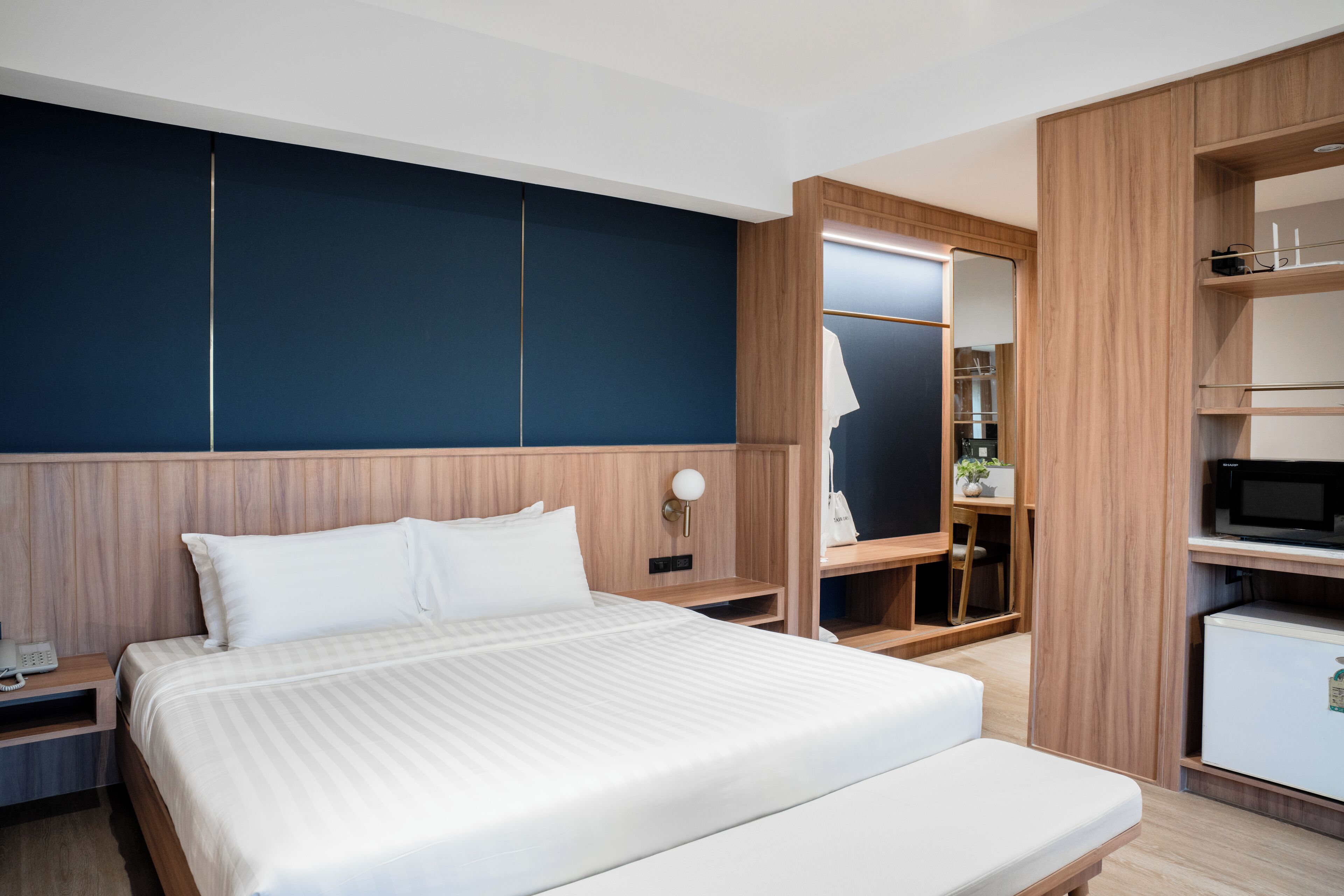A modern hotel room with a large bed, wooden paneling, a built-in wardrobe, a small kitchenette, and a bedside table with a lamp.