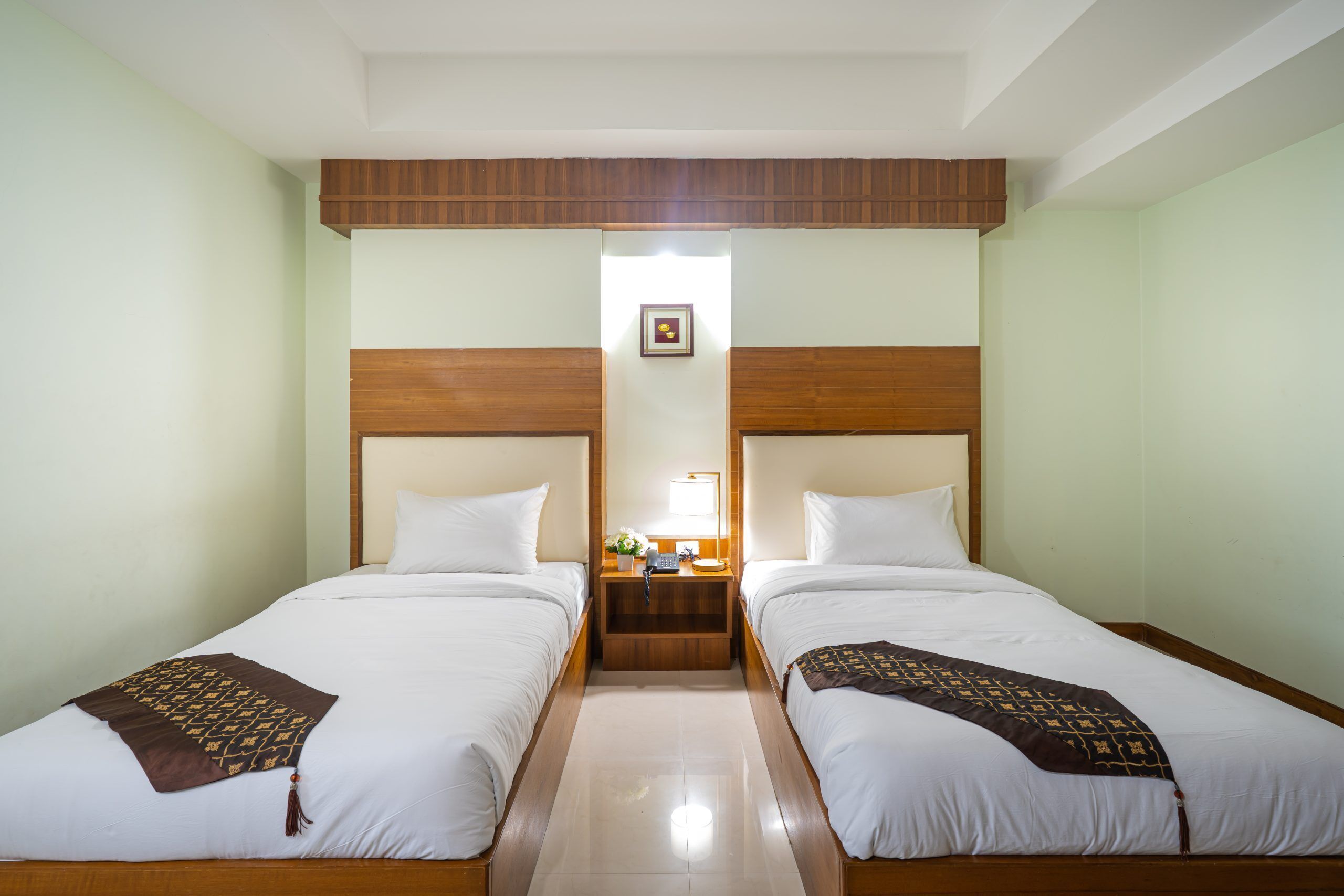 A hotel room with two single beds, wooden headboards, a nightstand with a lamp, and a framed picture on the wall.