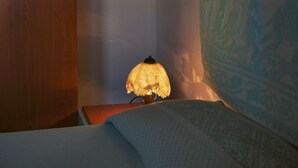 En säng med ett mönstrat överkast, en sänglampa med en lampskärm i spets och en vägg prydd med en väv.