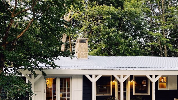 A house with a white exterior, a stone chimney, and a gravel patio with two wicker chairs and a table.
