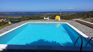 A rectangular swimming pool with a clear blue water surface, surrounded by stone tiles. There are two white lounge chairs with yellow covers, and a lamp post in the background.