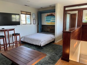 A bedroom with a bed, a desk with two chairs, a TV, and a mural on the wall.