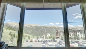 Vista desde una ventana sobre un estacionamiento con vehículos, un edificio de varios pisos y un paisaje montañoso al fondo.