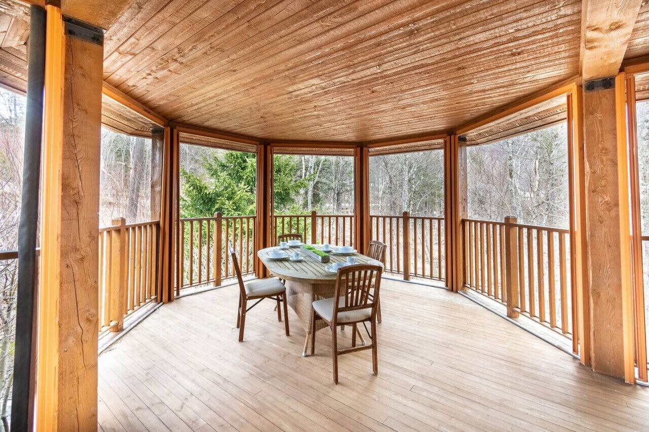 A covered outdoor dining area with a wooden table and chairs, surrounded by a wooden railing and large windows.