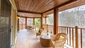 A covered outdoor area with wooden flooring, wicker furniture, and large windows offering a view of trees. - Tremblant Prestige - Panache 632 (Mont-Tremblant)