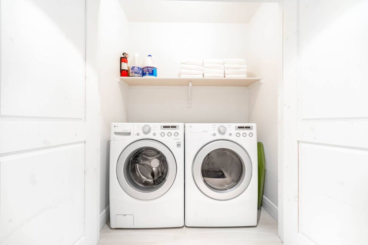 A laundry room with two white front-loading washing machines and a shelf above containing cleaning supplies.