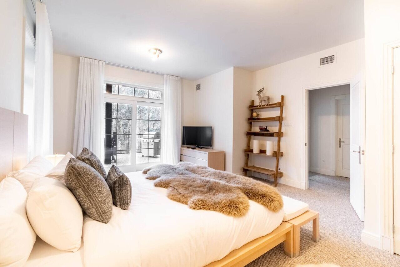 A bedroom with a bed, a television, a window with curtains, a shelf with decorative items, and a door.