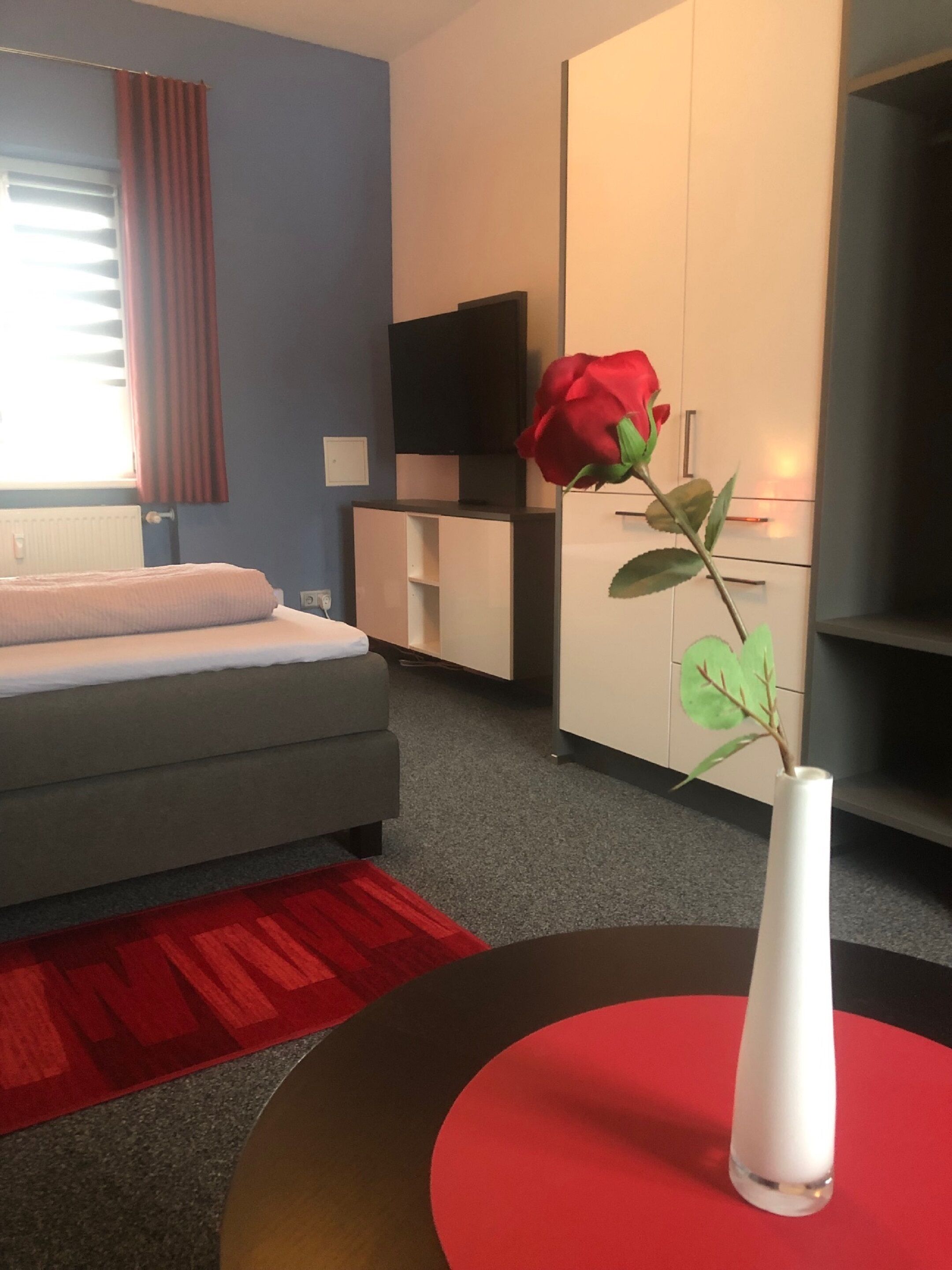 A modern hotel room with a grey sofa, a red accent rug, a white vase with a single red rose, and a flat-screen TV mounted on the wall.