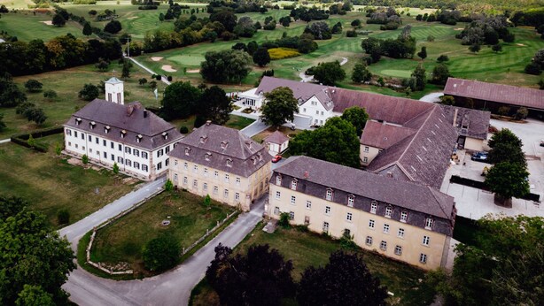 An aerial view of a large estate with multiple buildings, including a prominent white structure with a tower, surrounded by expansive green fields and a golf course. - Appartements am Rindhof (Muennerstadt)