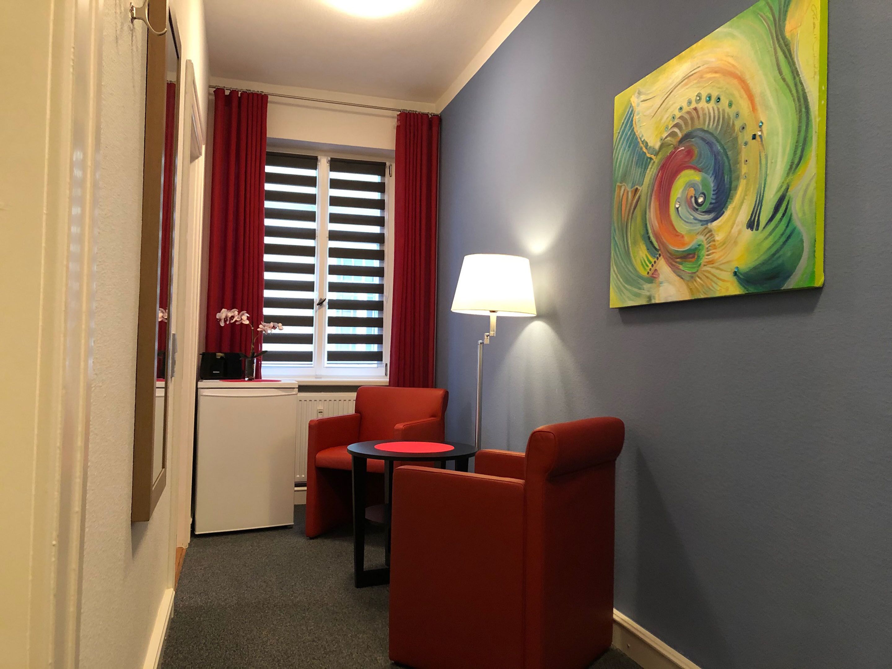 Ett litet rum med ett skrivbord, två röda stolar, ett litet bord, en lampa, ett fönster med röda gardiner och en färgstark, abstrakt målning på väggen.