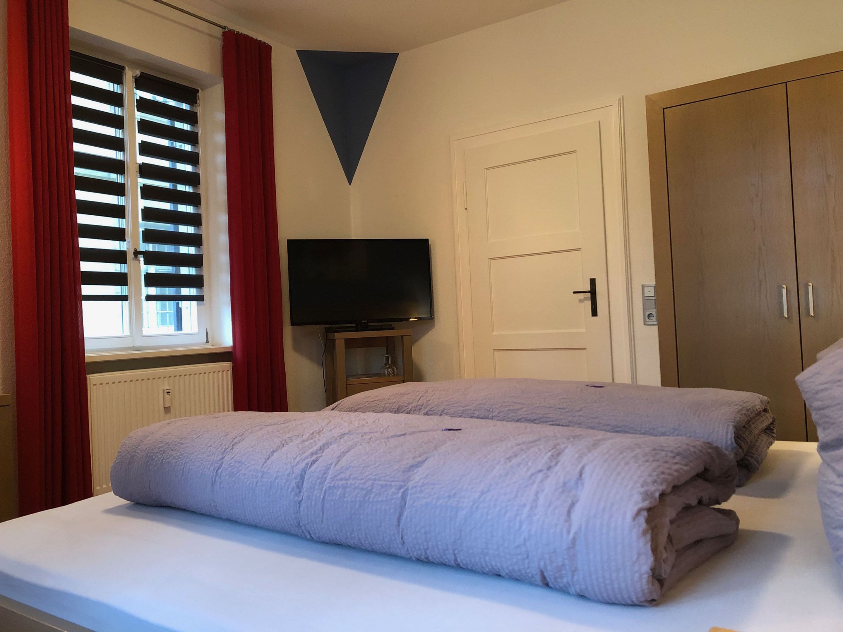 A bedroom with a bed, a television on a stand, a wardrobe, and a window with blinds.