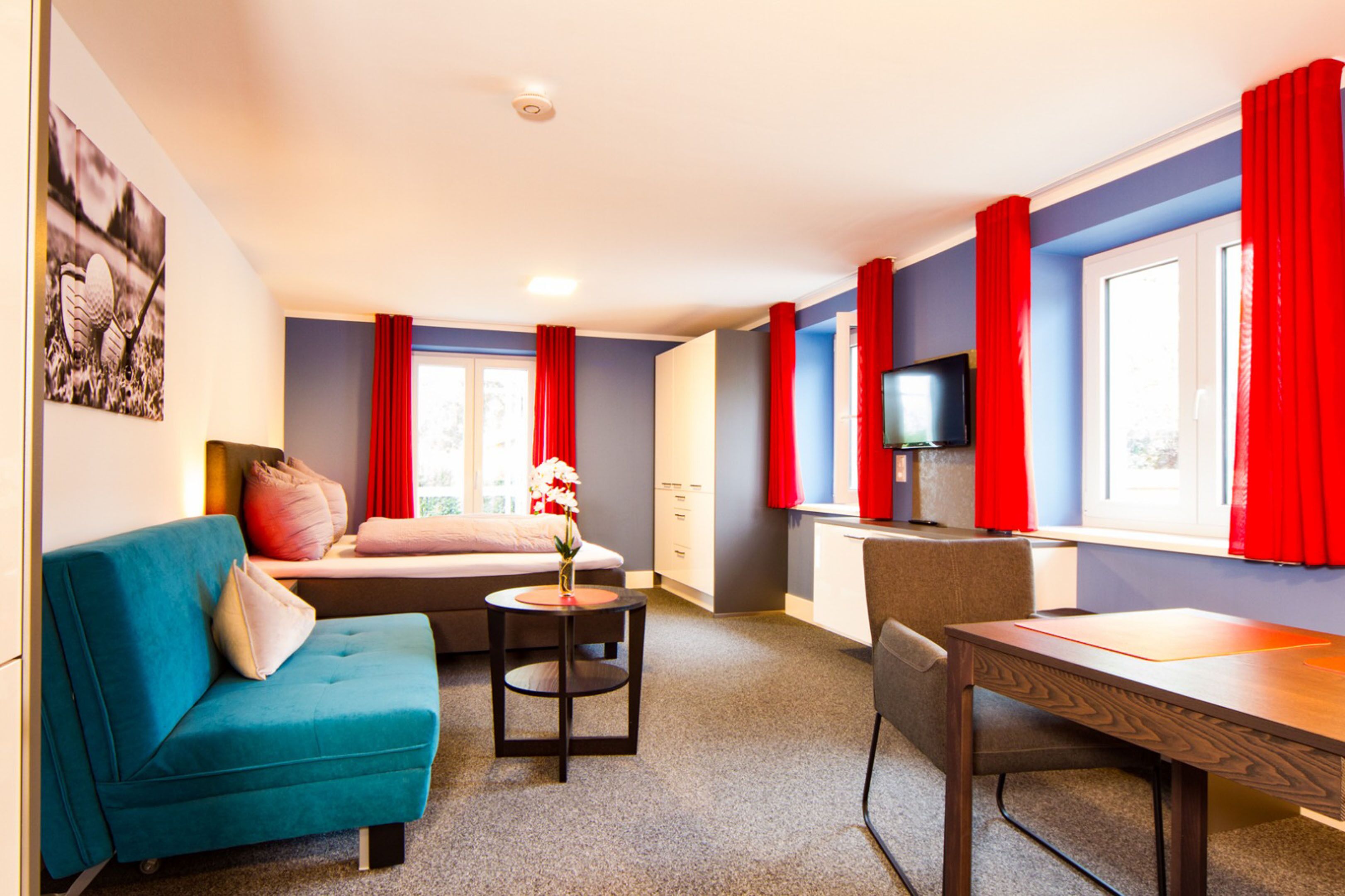 A room with a blue sofa, a small table with a vase, a bed with red curtains, a TV on a stand, and a window with red curtains.