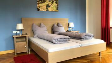 Un lit double avec une tête de lit en bois, deux tables de chevet avec des lampes, un tapis rouge et un tableau représentant un paysage accroché au mur.