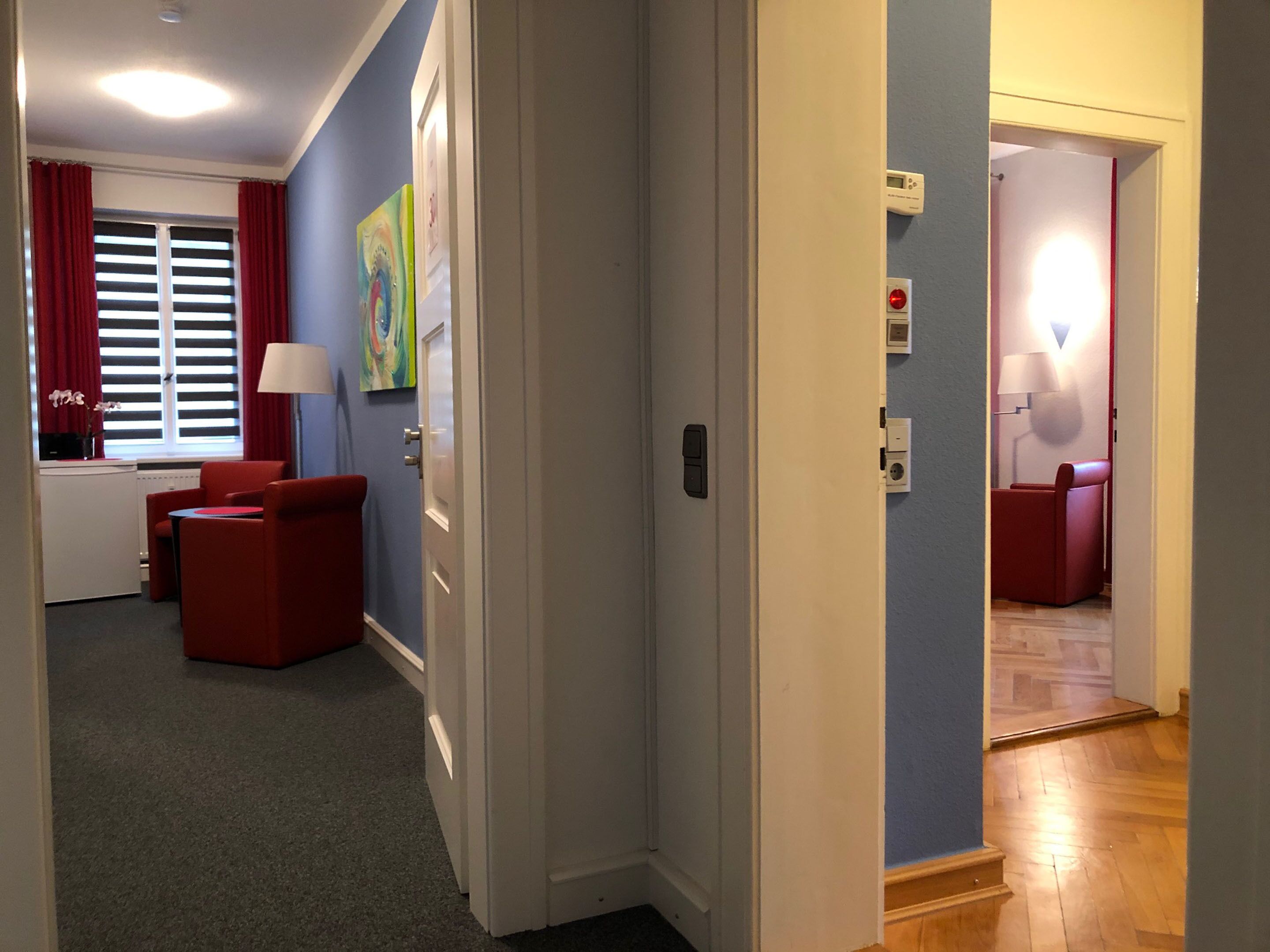 Quarto de hotel com sofá vermelho, uma mesinha, uma luminária e janela com cortinas vermelhas.