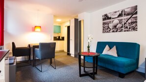 A modern living room with a teal sofa, a small round table, and a desk with chairs. A kitchen area is visible in the background. - Appartements am Rindhof (Muennerstadt)