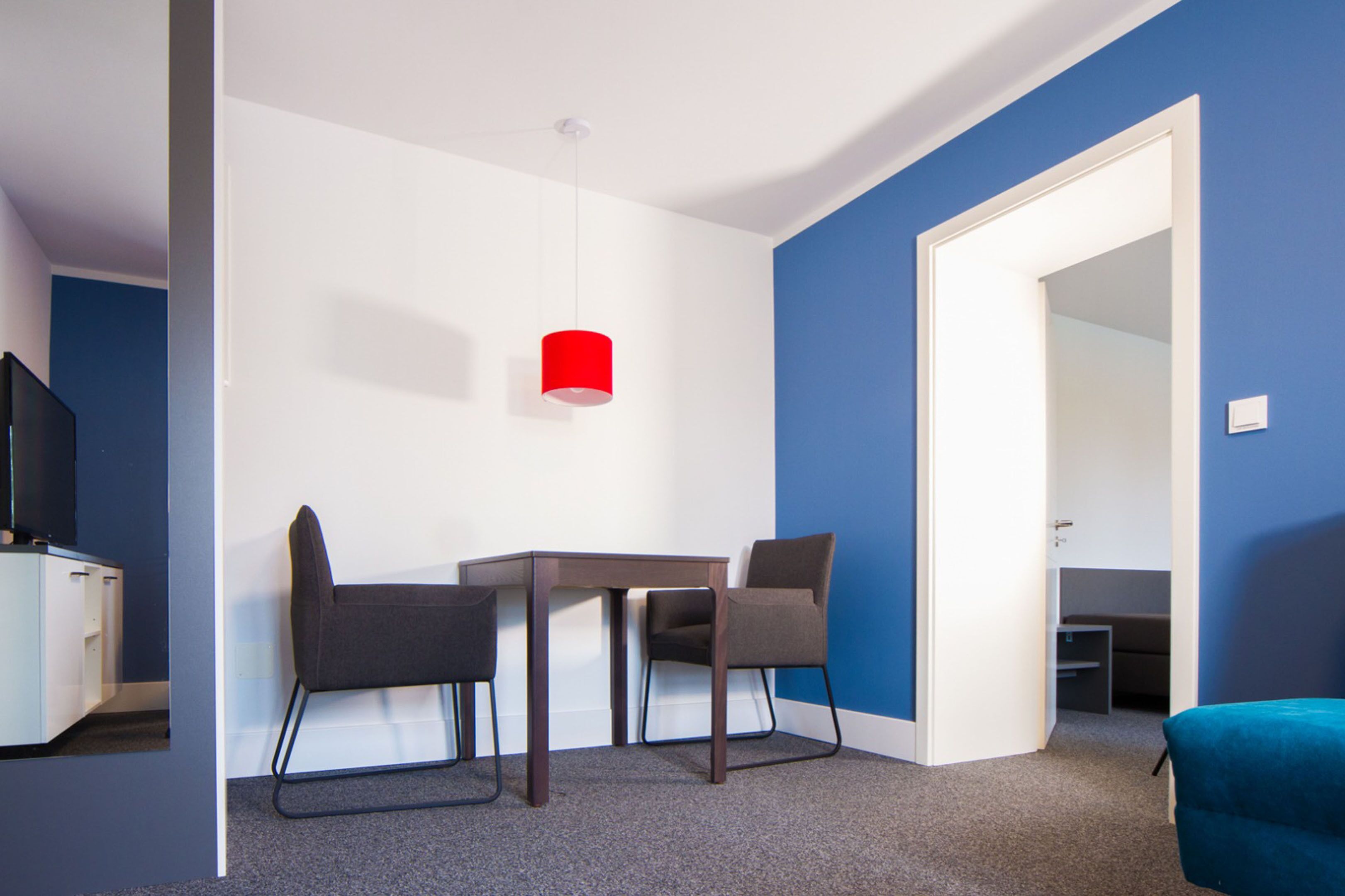 Ein Zimmer mit Schreibtisch, zwei Stühlen, einer roten Pendelleuchte, einem Fernseher und einer blauen Wand.