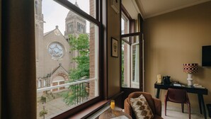 Una habitación con un ventanal grande que da a un edificio histórico con una torre de reloj. Hay una silla con cojín, una mesita con un periódico y un vaso de jugo, y un escritorio con una lámpara y un libro.