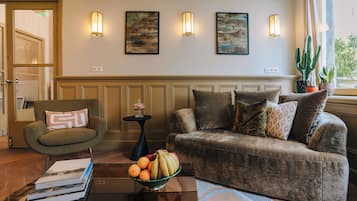 Un salón con un sillón verde, un sofá marrón y una mesa de centro de vidrio con libros y fruta.