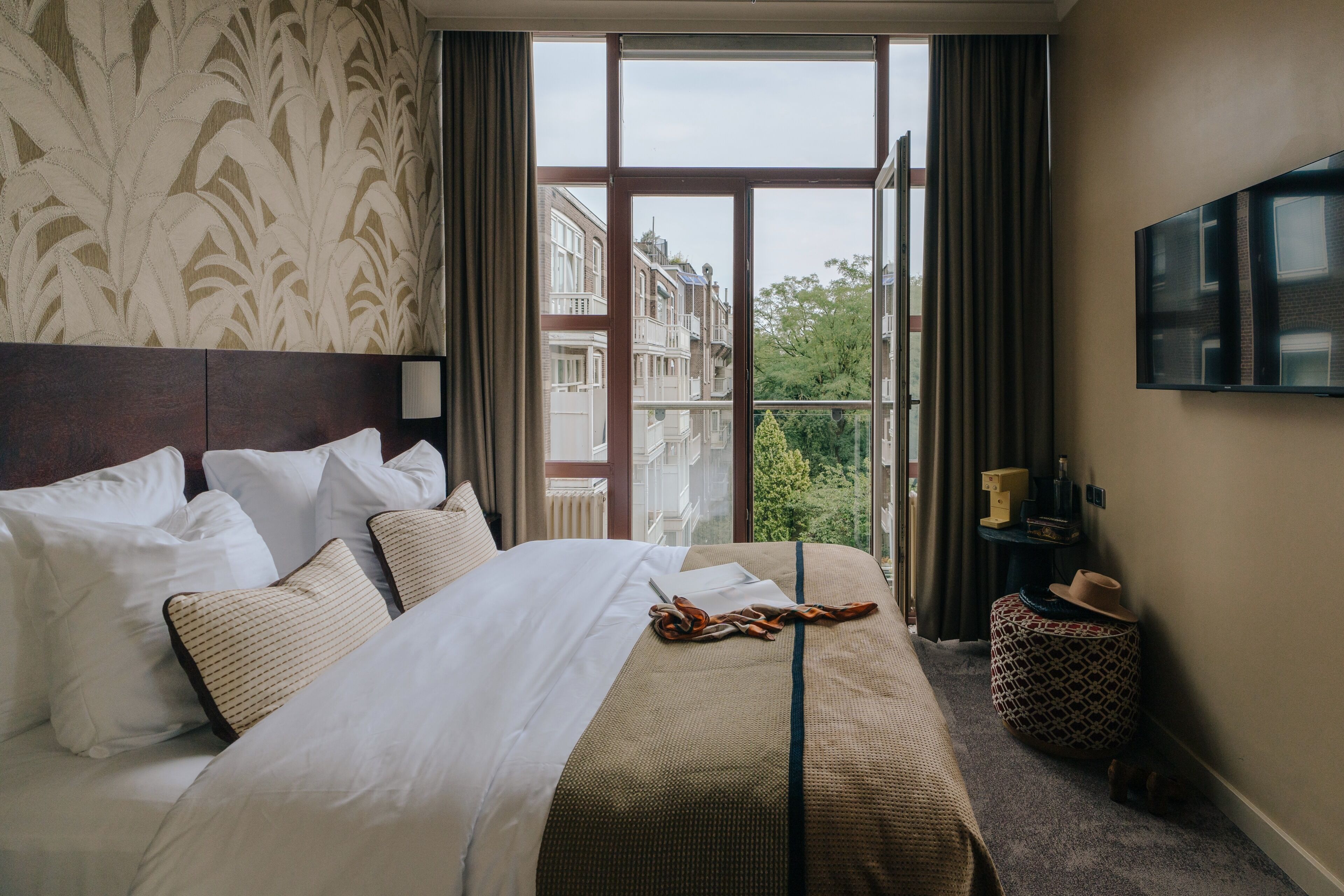 Un cuarto de hotel con una cama grande, un televisor montado en la pared y vistas de edificios a través de la ventana.