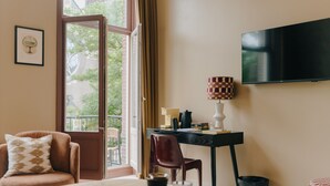 Habitación de hotel con cama, escritorio, silla, televisor y ventana con vistas a edificios y árboles.