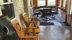 A rustic interior with wooden walls, a stone accent wall, a dining area with wooden chairs and a table, and large windows offering a view of the outdoors. - Rustic Lodge near Black Moshannon & Penn State (Philipsburg)