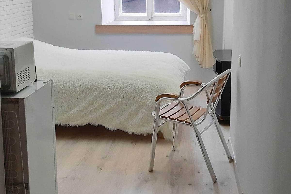 Una habitación pequeña y luminosa con una cama, una silla y un ventanal con cortinas.