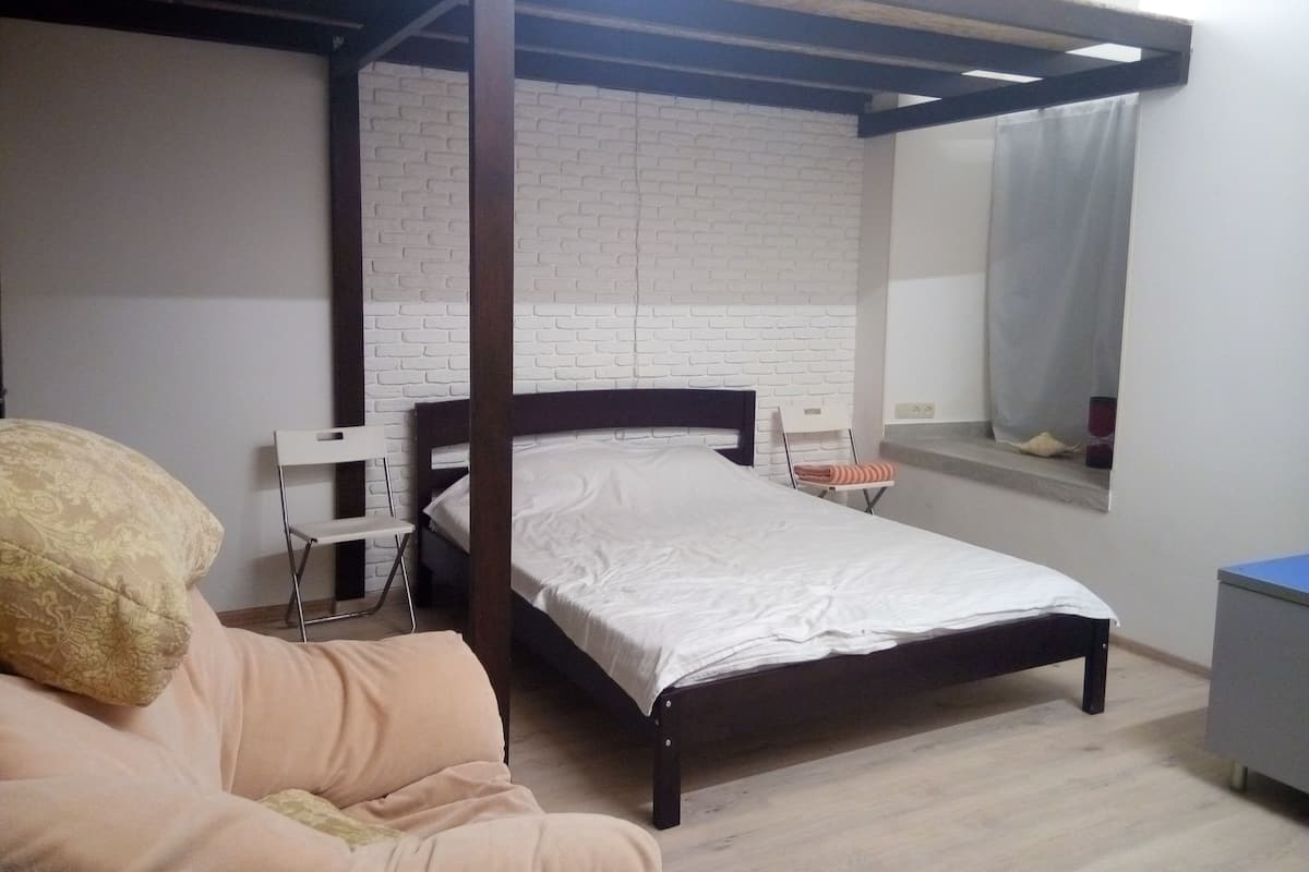 Un dormitorio con cama, una silla, una mesita y una ventana con cortina.