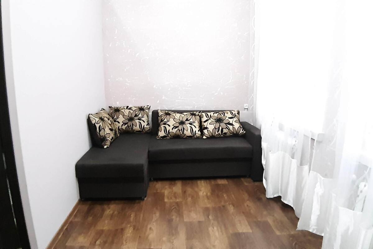 Una habitación de esquina con un sofá seccional negro, dos almohadas a juego y una cortina blanca.