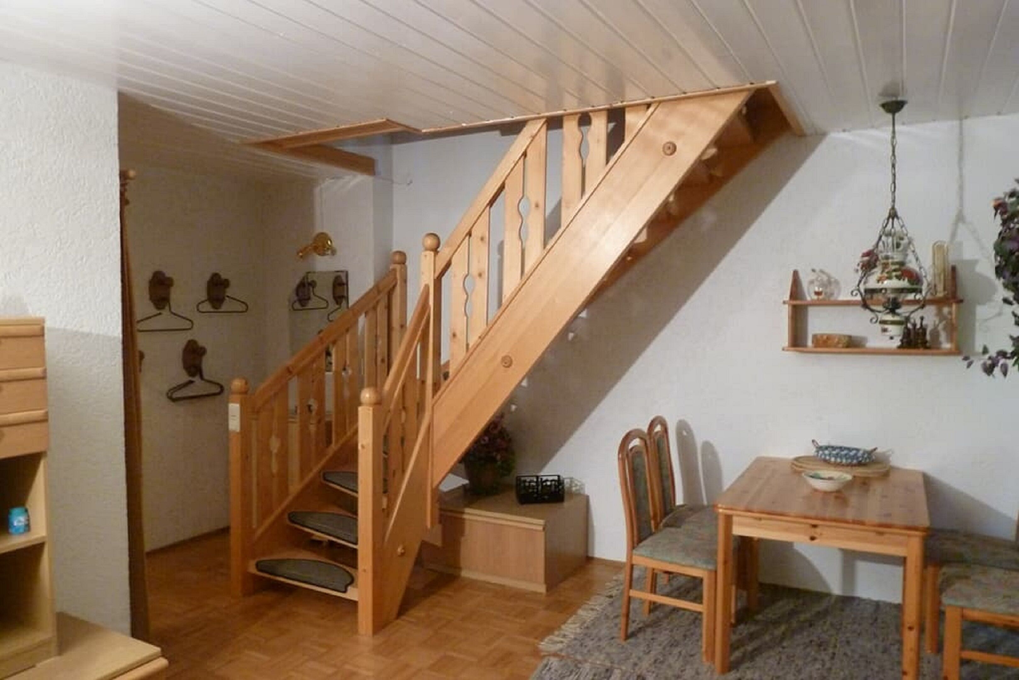 Eine Holztreppe führt zu einem Erkerbereich mit einem Esstisch, Stühlen, einer Kandelaberleuchte und Wandregalen.