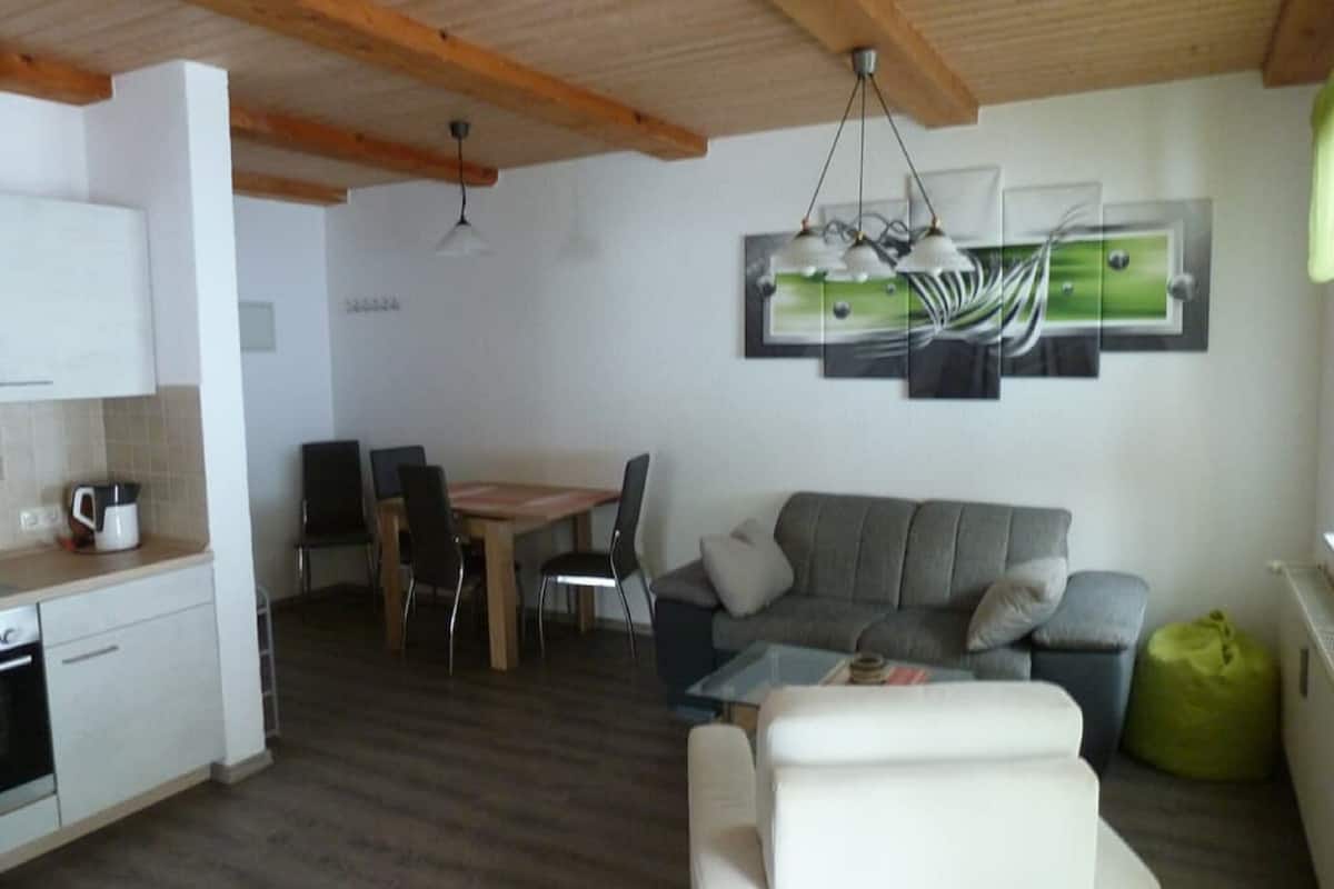 Une cuisine moderne avec des meubles blancs, un coin repas avec une table et des chaises en bois, un canapé gris et un fauteuil en forme de haricot vert.