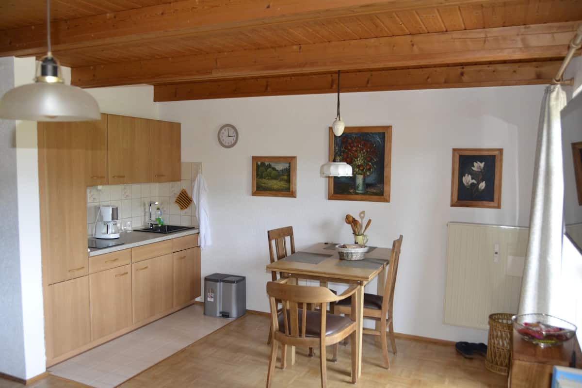 Une cuisine avec des meubles de rangement en bois, une table à manger avec des chaises, une horloge et un tableau accroché au mur.