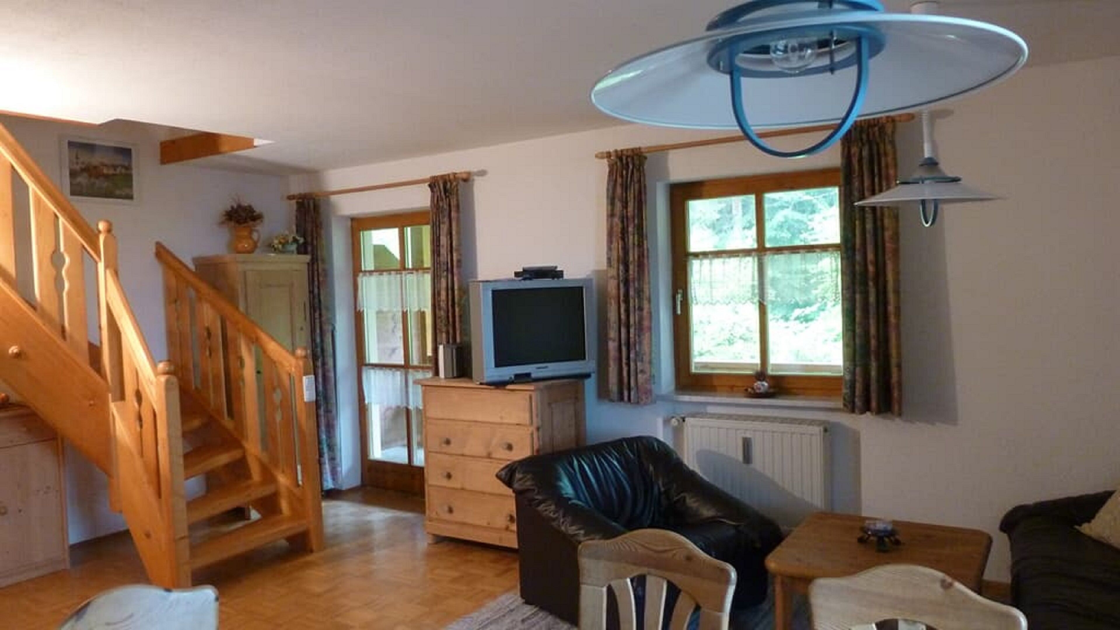 Ein gemütliches Wohnzimmer mit einer Holztreppe, einem Fernseher auf einer Kommode, einem schwarzen Ledersofa und einem Essbereich mit Tisch und Stühlen.
