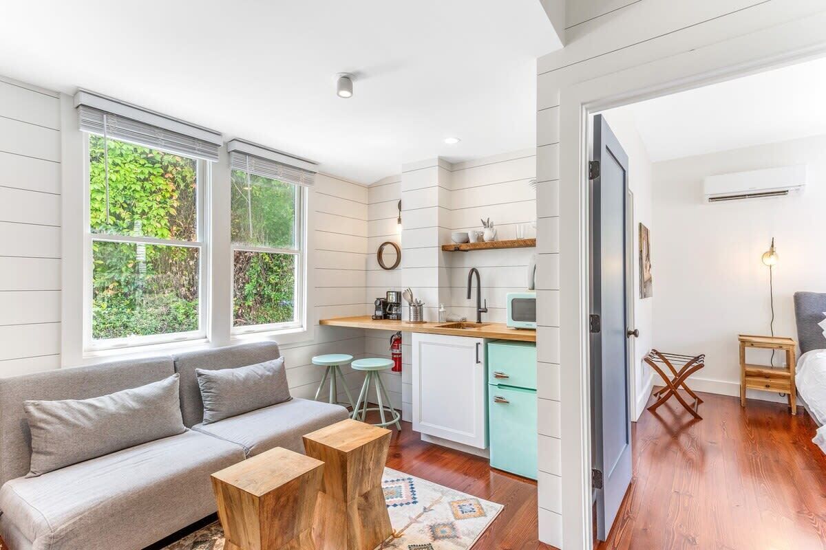 Een compact woongedeelte met een kitchenette, een bank, een salontafel en uitzicht op groen via grote ramen.