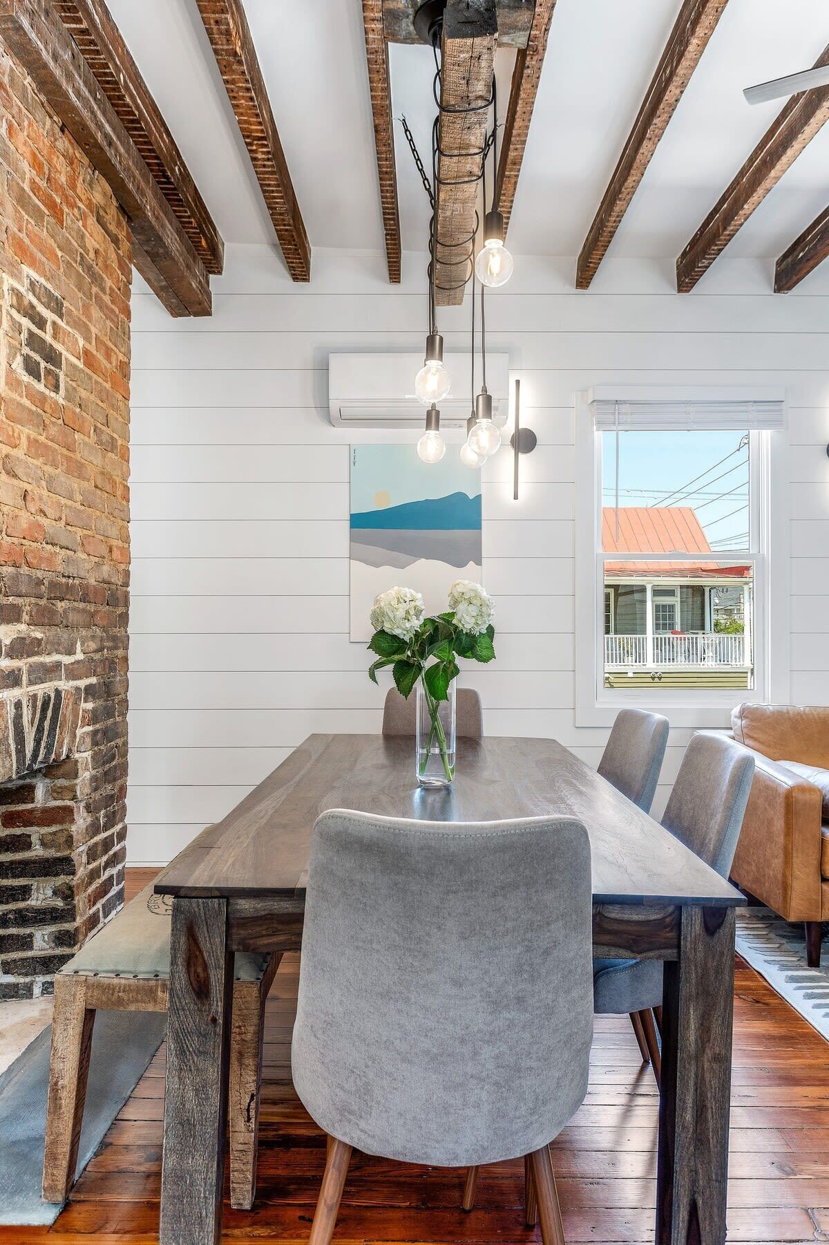 A dining area with a wooden table, grey upholstered chairs, a vase with white flowers, and exposed brick walls.