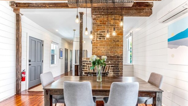 A dining area with a wooden table, grey upholstered chairs, a brick wall, hanging light bulbs, and a fire extinguisher on the wall. - The Sunrise | 3BR Up - Best of Charleston Award! (Charleston)