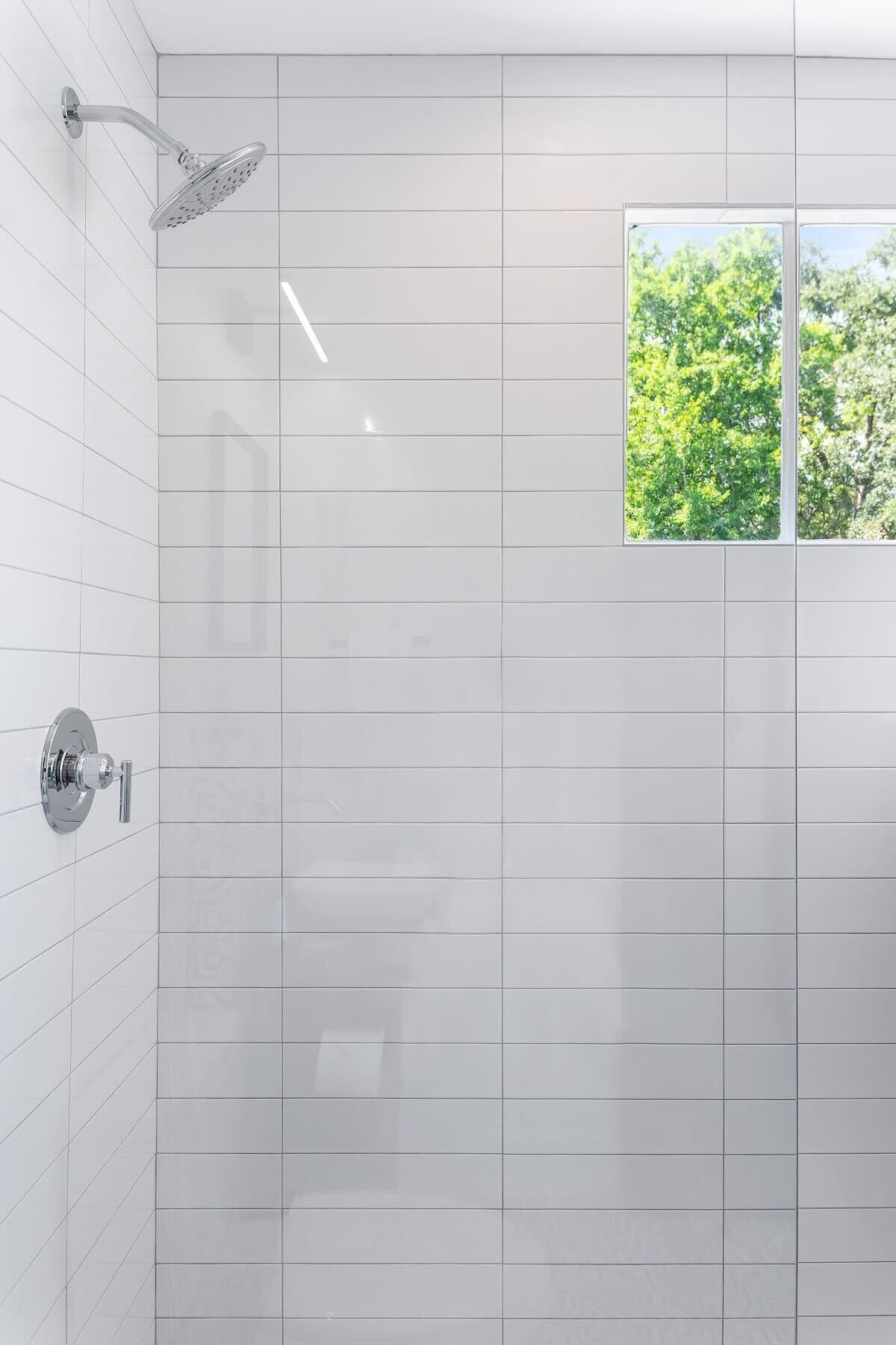 Un baño moderno con azulejos blancos, un cabezal de ducha y una ventana con vistas a la vegetación.