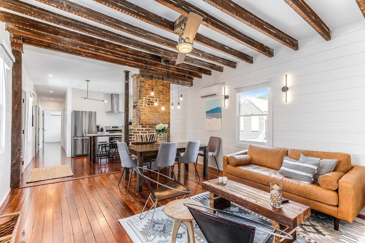 A spacious living area with a wooden floor, a leather sofa, a dining table with chairs, and a kitchen in the background.