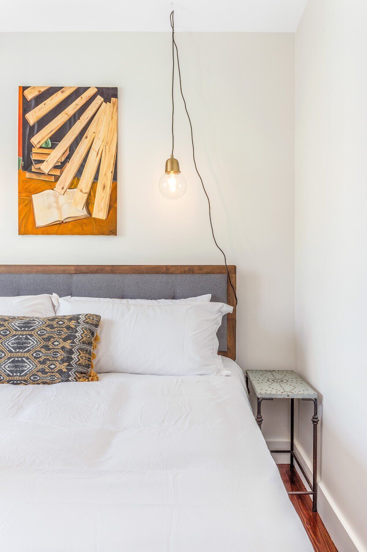A bedroom with a bed, a bedside table, a wall-mounted light, and a painting.