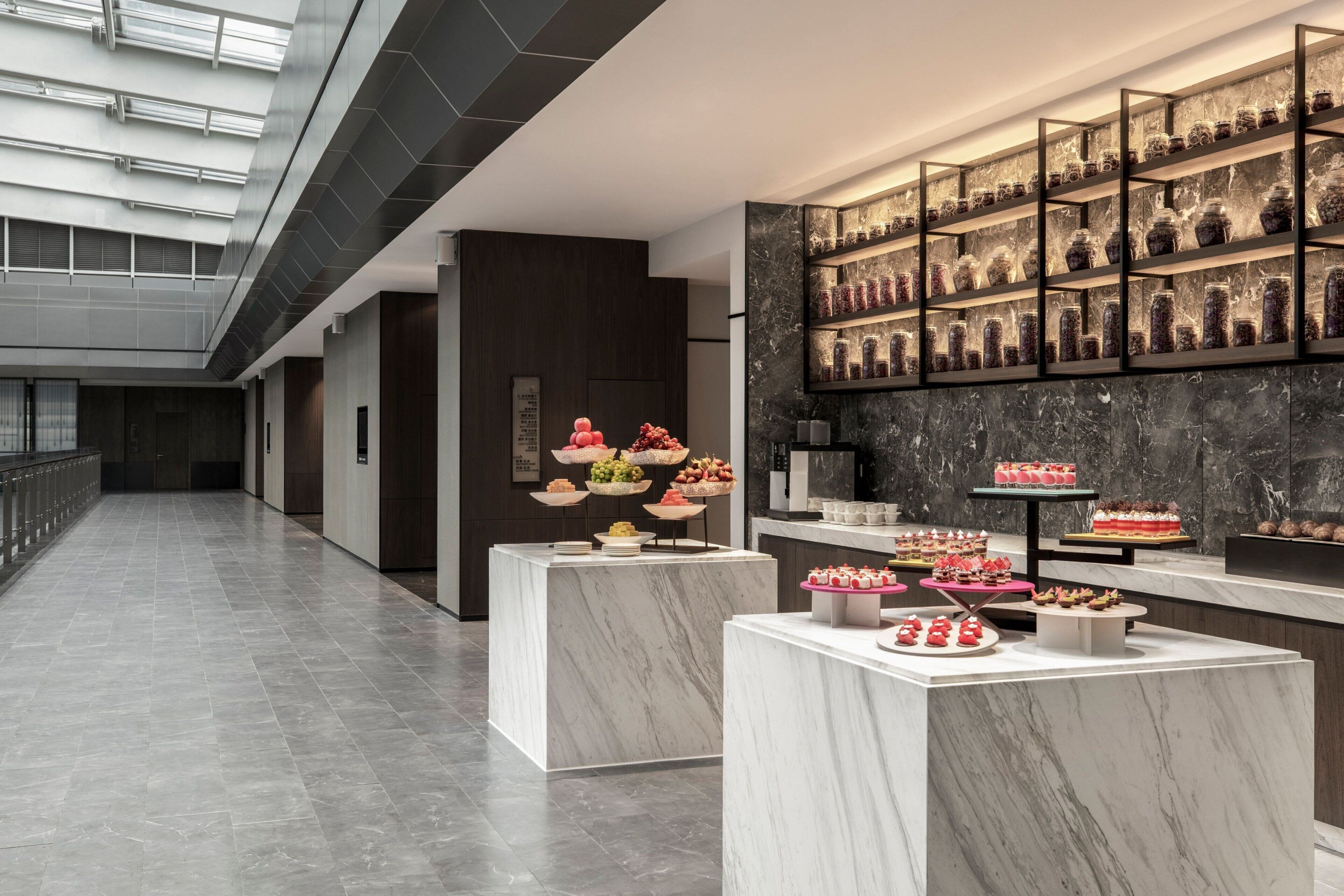 A modern hotel lobby with marble flooring, a glass ceiling, and a wall displaying various food items.