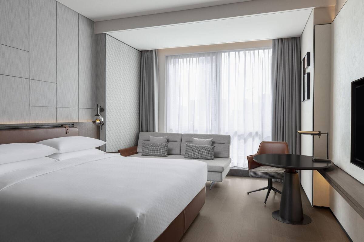A modern hotel room with a large bed, a sofa, a dining table, and a TV.
