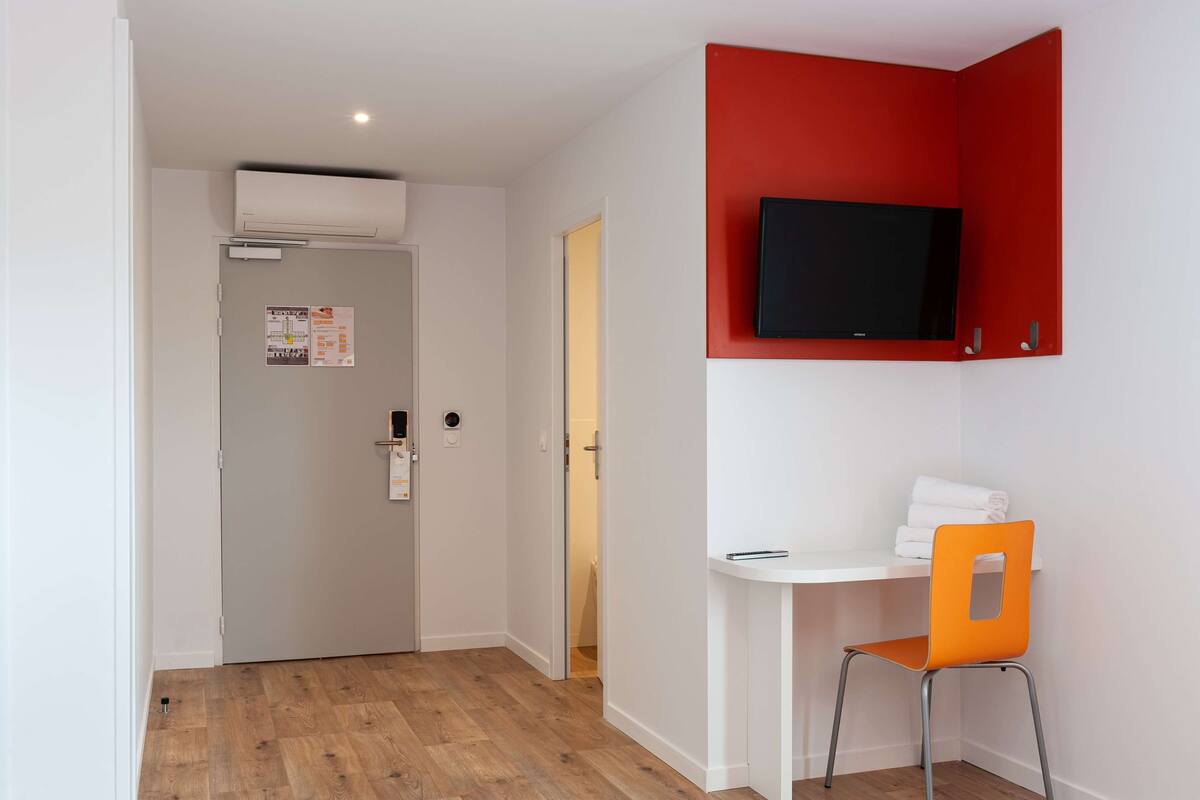 A room with a white desk, an orange chair, a red accent wall, a mounted TV, and an air conditioning unit.