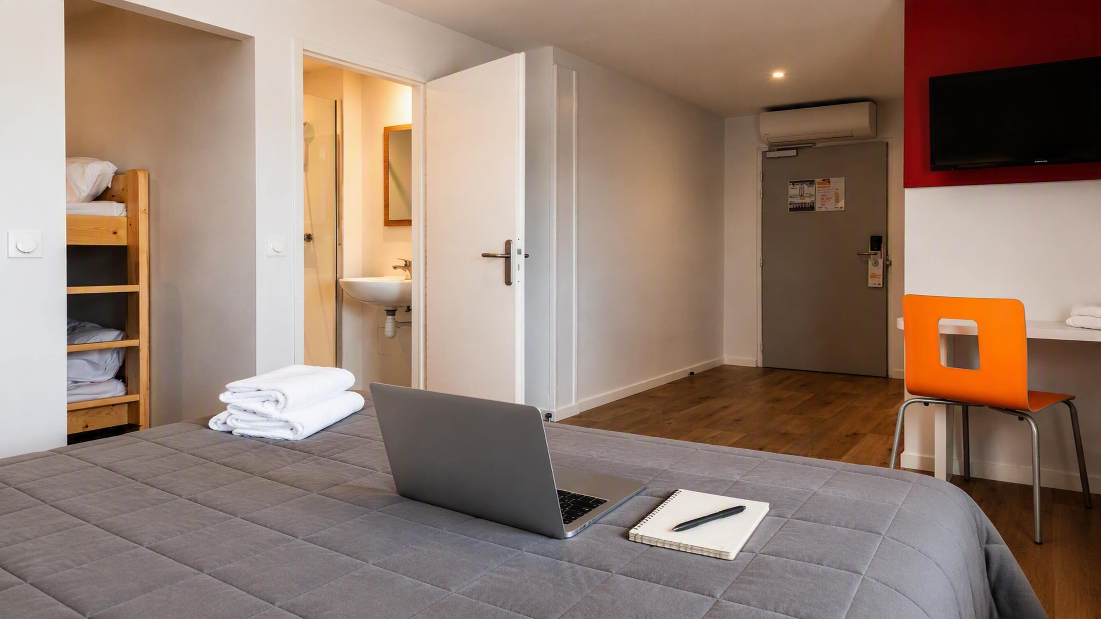 A bedroom with a bed, a laptop, a notebook, a chair, a wardrobe, a bathroom, and a TV.