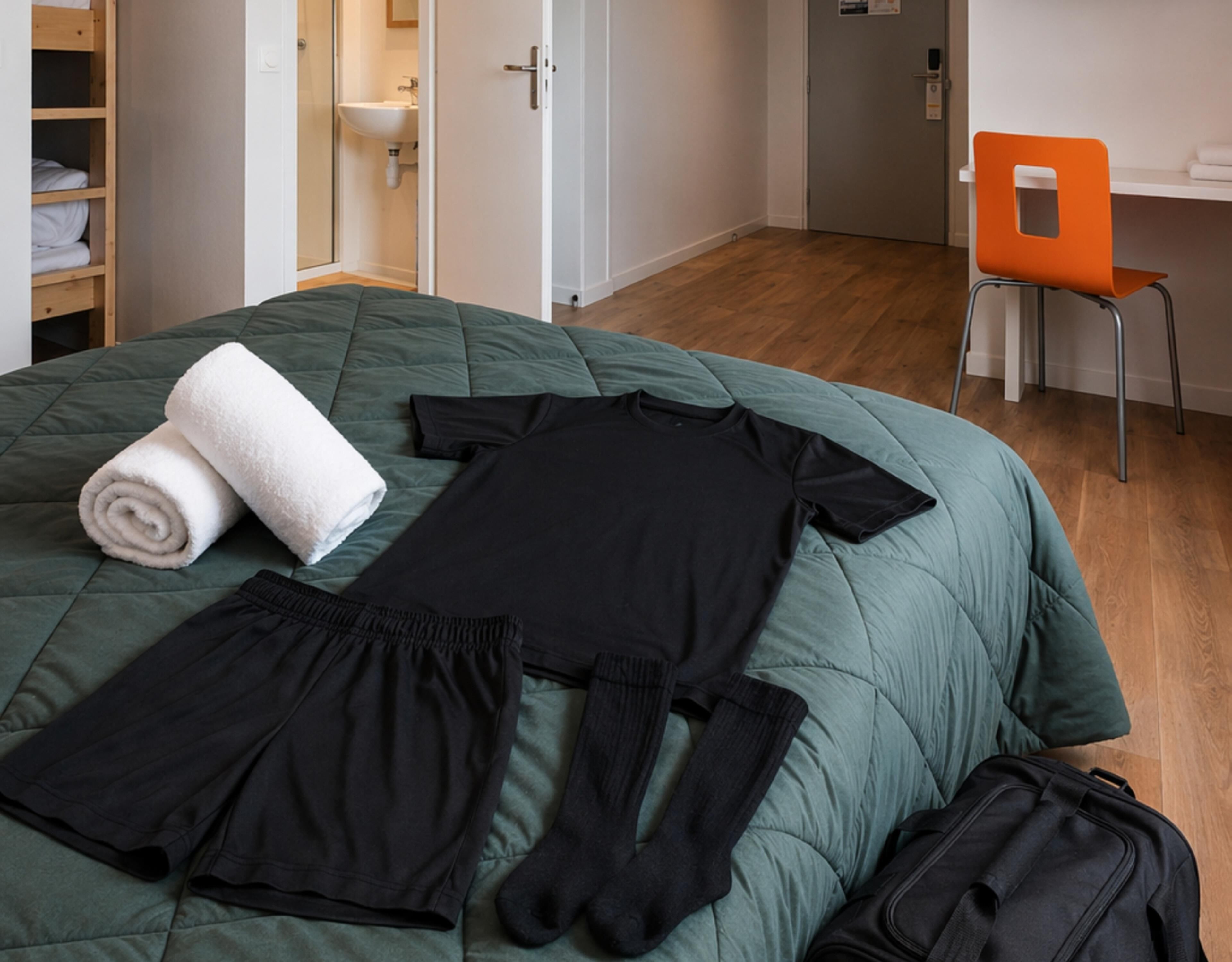 A neatly made bed with a green duvet, a rolled white towel, black clothing, and a black suitcase on a wooden floor.
