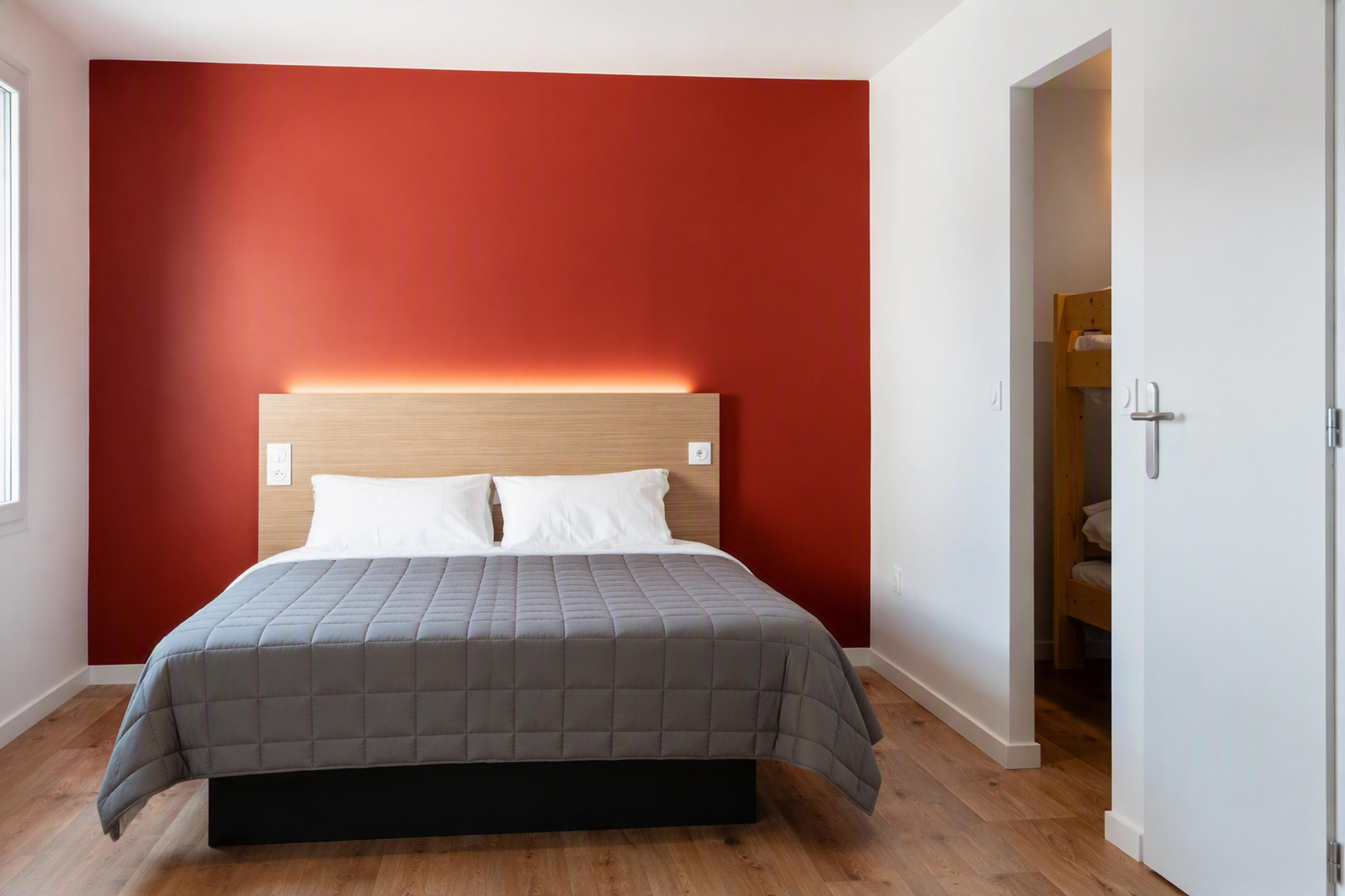 A bedroom with a bed, a red wall, and a wooden floor.