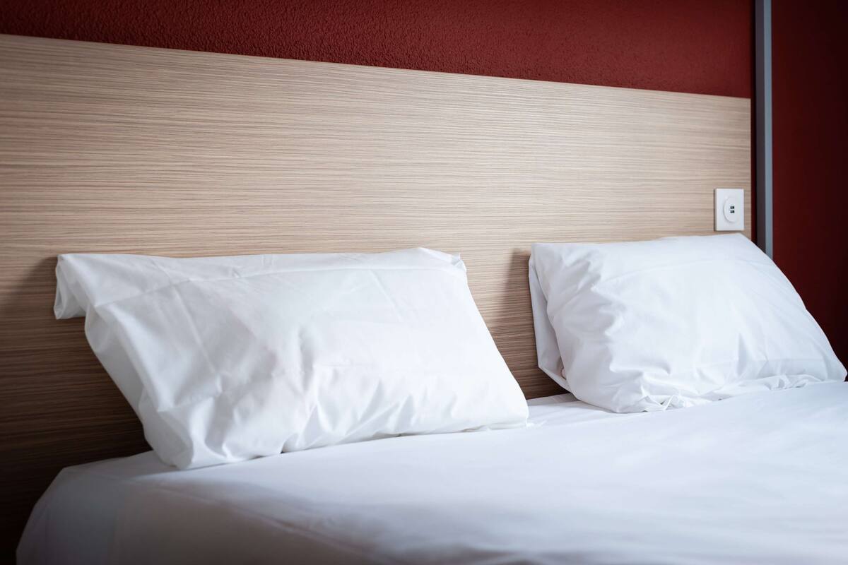 A neatly made bed with white linens and pillows, a wooden headboard, and a red wall.