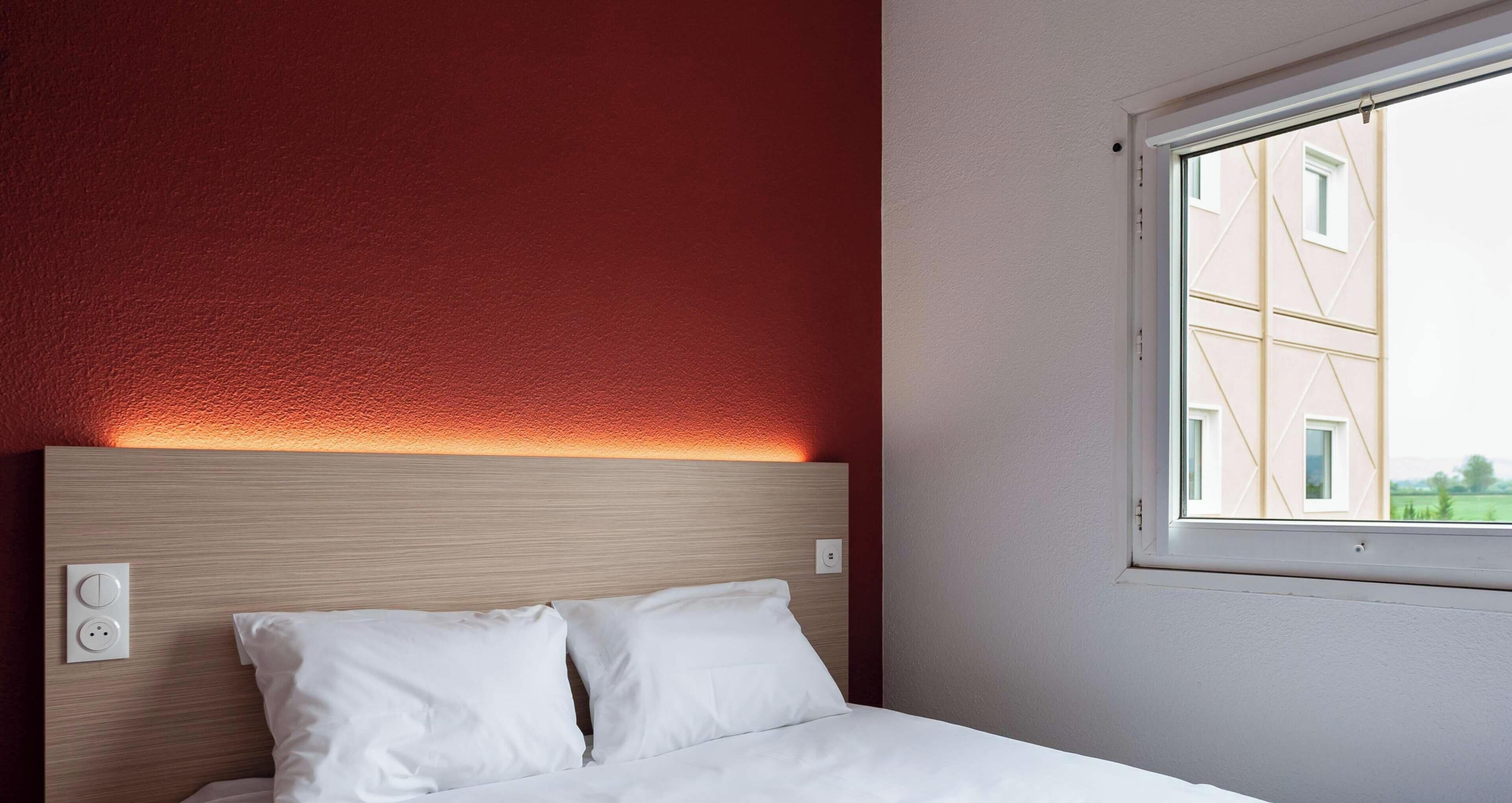 A hotel room with a bed, a headboard, a window, and a wall socket.