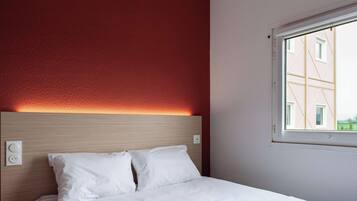 A hotel room with a bed, a headboard, a window, and a wall socket.