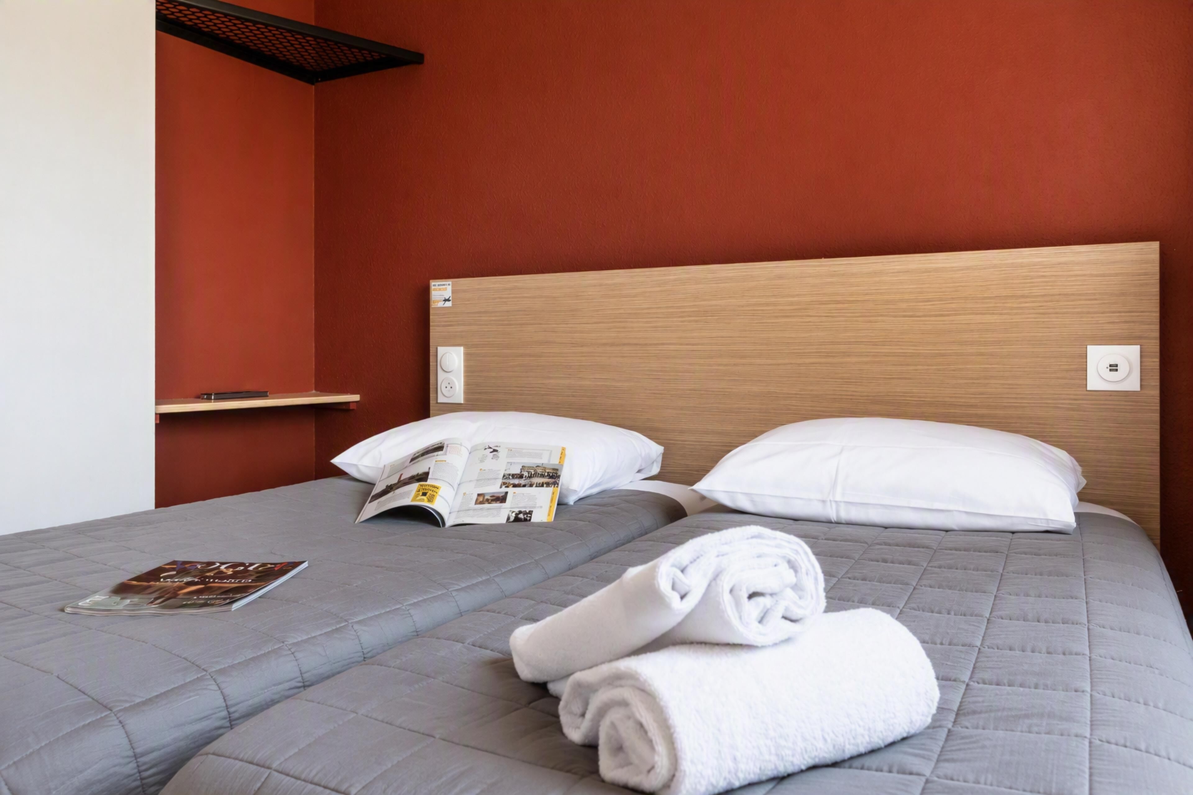 A hotel room with a single bed, a bedside table, a magazine, and a rolled towel.